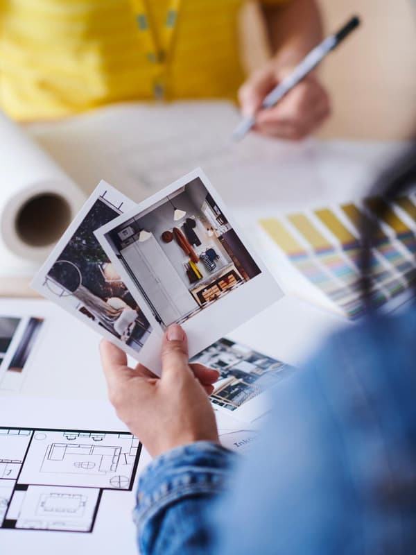Person holding two photos, one showing an interior; in the background, color swatches and plans on a table.