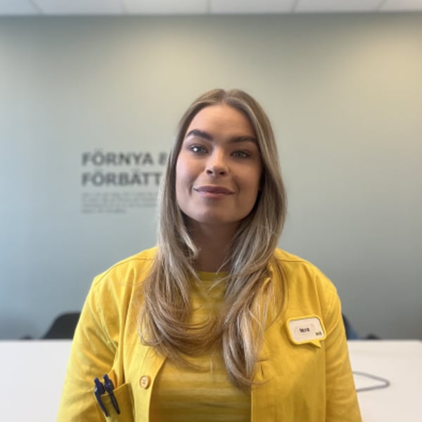 Person i gul arbetsuniform med långt ljust hår står i ett ljust kontorsrum med text på väggen i bakgrunden.