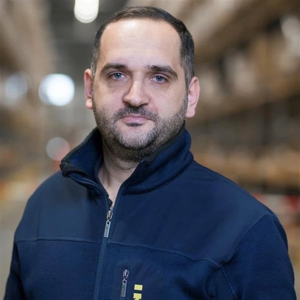 Person i mörkblå arbetsjacka står i en lagermiljö med hyllor och oskarpa ljuskällor i bakgrunden.