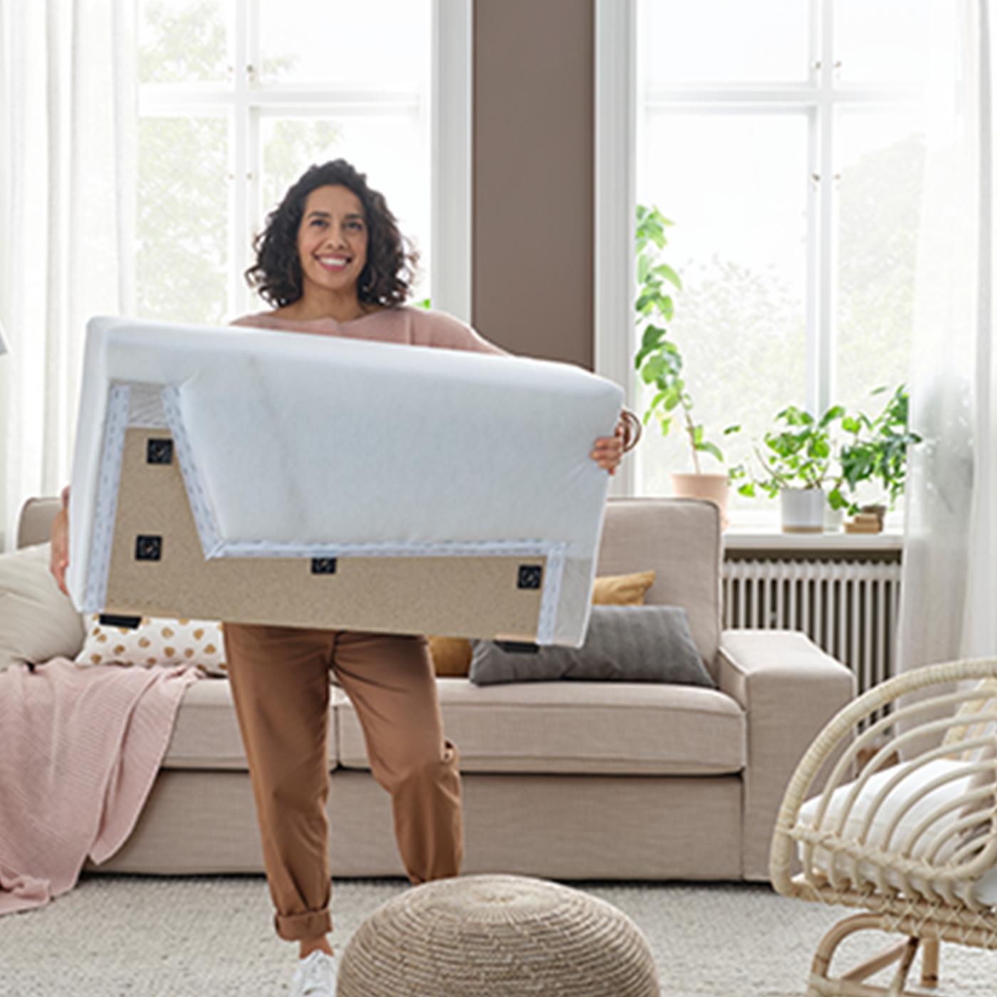 Person i vardagsrum som håller i ett stort vitt rektangulärt föremål, troligen en möbel eller dyna. Personen har bruna byxor och vita skor. Rummet har en beige soffa med kuddar, växter i fönstret, stora fönster med naturligt ljus och en korgstol i förgrunden.