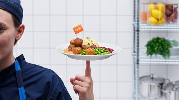 Person in dunkler Kochkleidung balanciert einen weißen Teller mit Fleisch-Gemüse-Bällchen, Erbsen, Kartoffelpüree und Preiselbeeren auf einem Finger. Auf dem Teller steckt ein kleines orangefarbenes Fähnchen mit der Aufschrift ‚New‘. Hintergrund: weiße Fliesenwand und Küchenregal.
