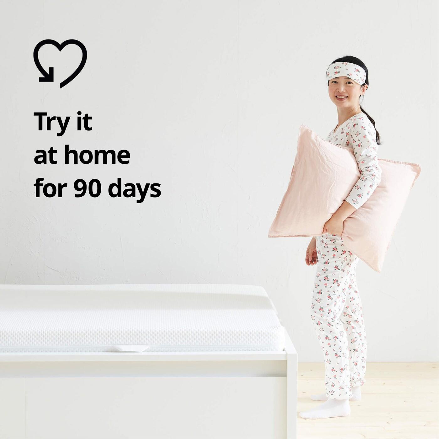 Person in pajamas next to bed with mattress, holding pink pillow; on left is text "Try it at home for 90 days" with heart-shaped symbol.