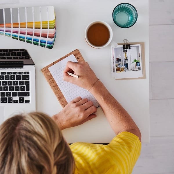 Person in yellow shirt planning home design with notepad, laptop, coffee, and color samples.