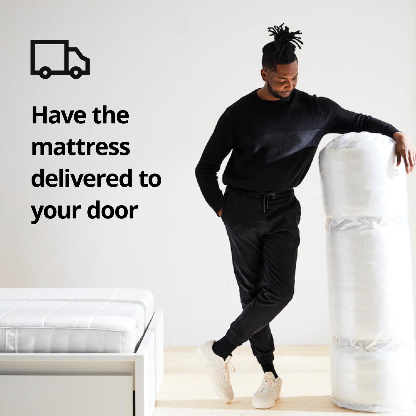 Person leaning against rolled up mattress in white room; on the left a bed with mattress and on the image the text "Have the mattress delivered to your door" with truck icon.