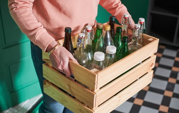 Person moving through a hall carrying a KNAGGLIG wooden crate filled with a variety of glass and plastic bottles.
