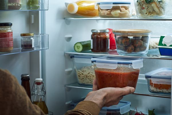 Person placing a glass IKEA 365+ food container filled with tomato sauce into a well-stocked fridge.