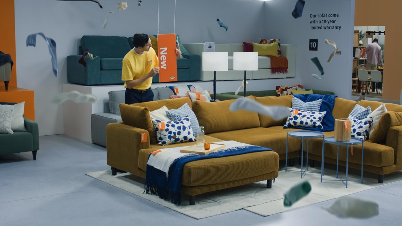 Person placing a "New" sign in an IKEA sofa display featuring sectional seating and accessories.