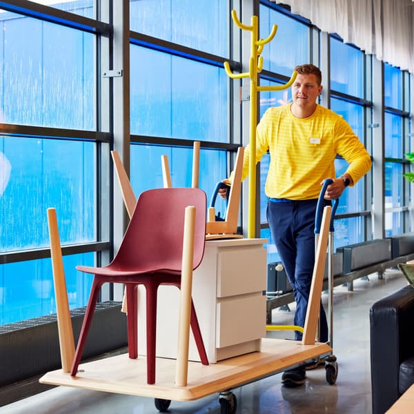 Person pushes cart with red chair, white drawers, and yellow coat rack near large blue-tinted windows indoors.