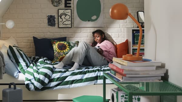 Person reclining on a striped bed, reading a handheld device in a cozy bedroom with colorful pillows and books.