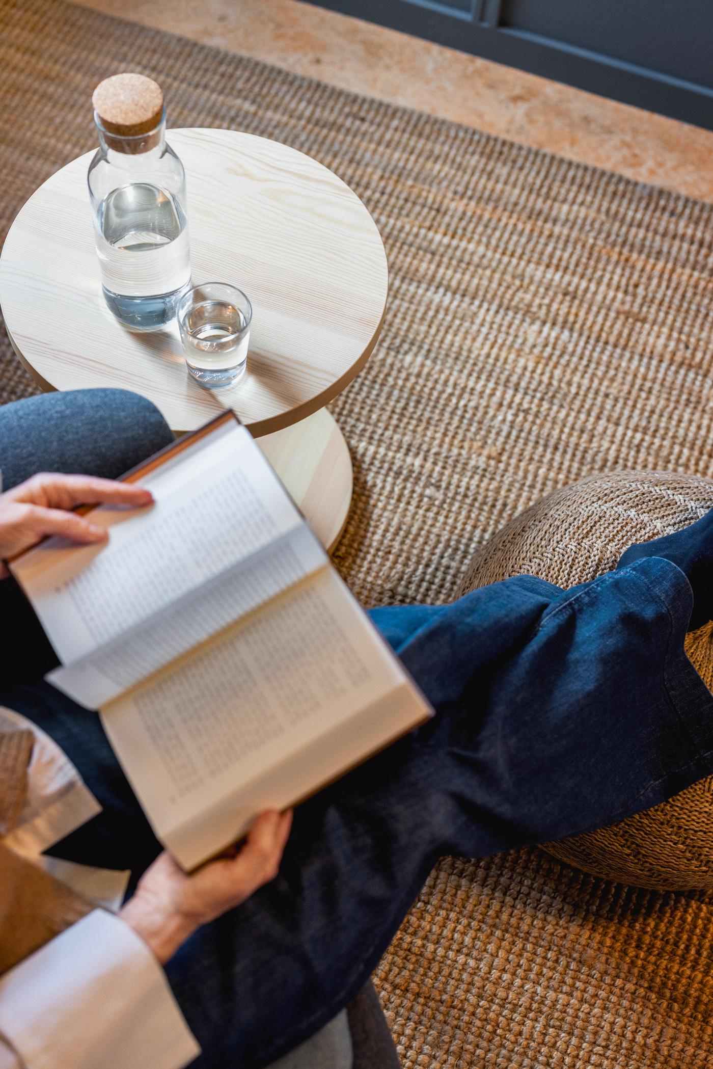 Person sitter med korslagda ben på en vävd matta och läser en bok, bredvid ett litet runt träbord med vattenflaska och glas.
