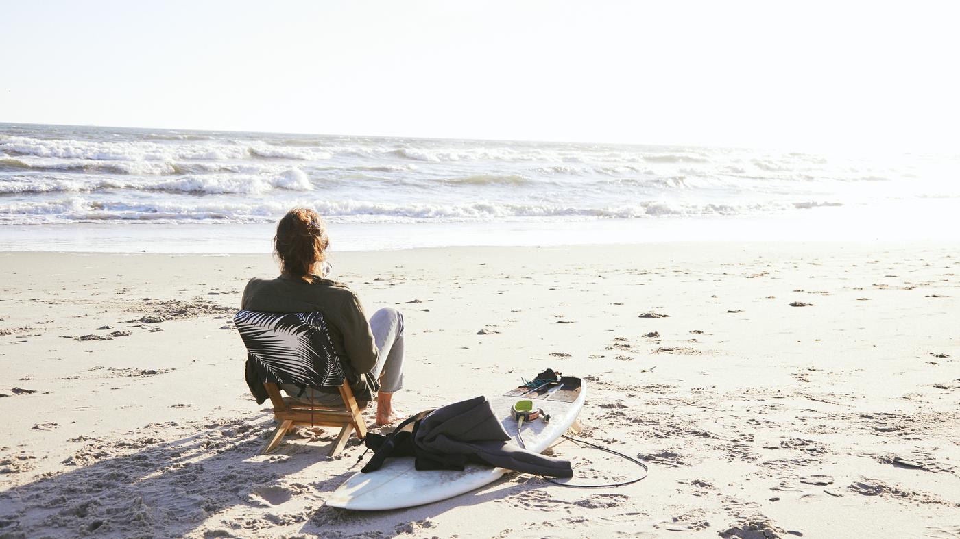 Person sitzt entspannt am Strand und blickt aufs Meer, neben einem Surfbrett und Neoprenanzug.