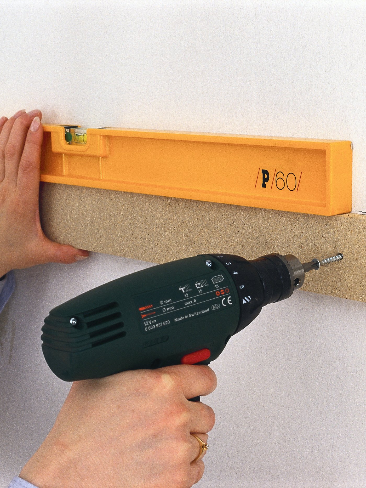 Person using a drill to mount a board on a wall while holding a level to ensure it is straight.