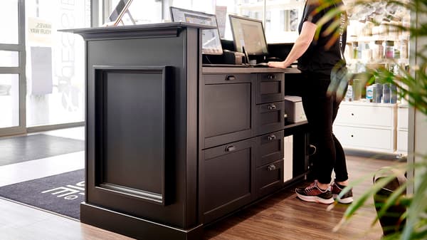 Person vid svart receptionsdisk med två skärmar, stora fönster bakom och produkter på hyllor till höger.


