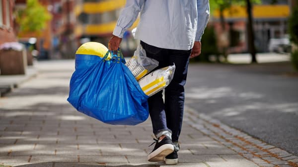 Person walking on sidewalk carrying a large blue shopping bag filled with household items.