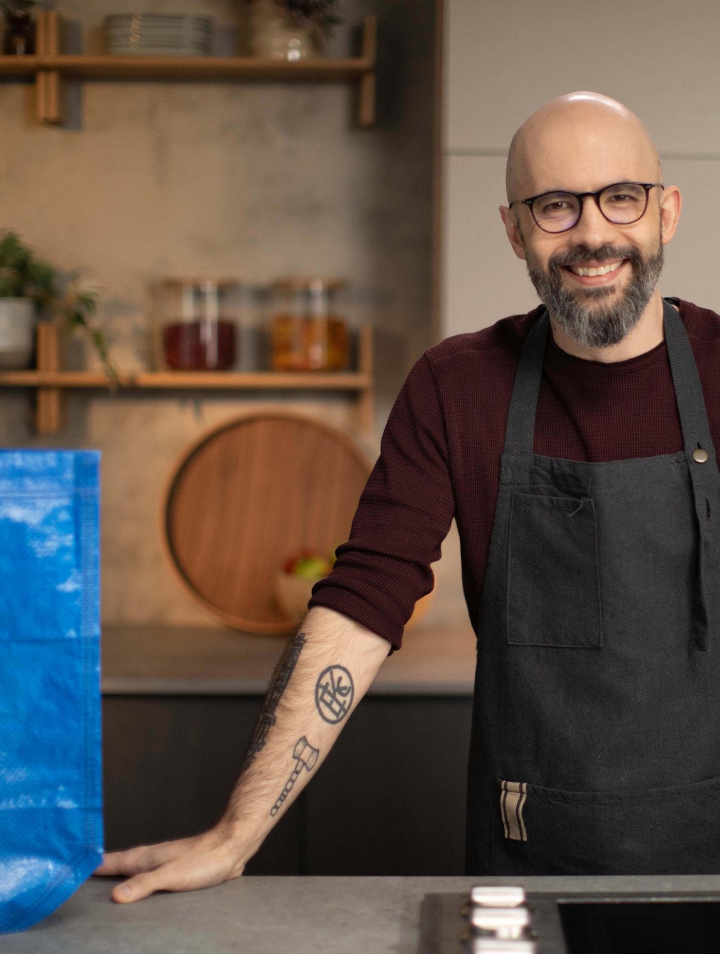 Person wearing an apron standing at a kitchen counter with shelves and a blue bag nearby.