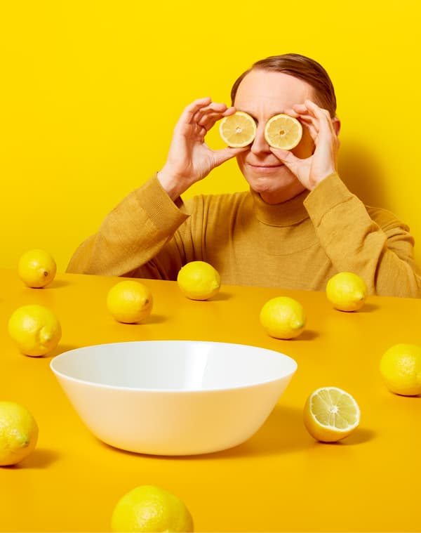 Person wearing yellow holding lemon slices over their eyes next to a yellow table with whole lemons and a white bowl on it.
