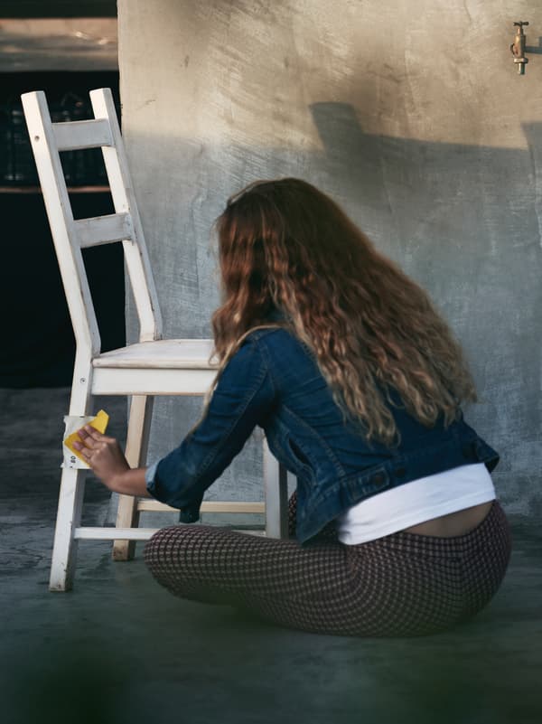 Person with long brown hair sitting on the floor in a yard sanding a white-painted wooden chair with sandpaper in sunlight.