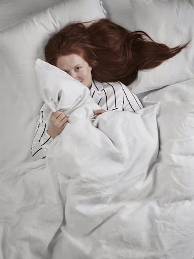 Person with long red hair lying in bed, partially covered by a white blanket, holding a pillow close to their face; wearing a striped shirt with white bedding.