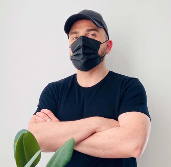 Persona amb gorra negra, mascareta negra i samarreta negra, amb els braços creuats davant d’una paret blanca i una planta al primer pla.