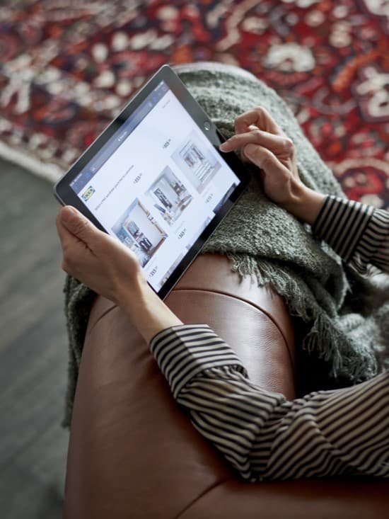 Persona asseguda en un sofà mirant una botiga en línia des d'una tauleta. A la pantalla es veuen mobles amb els preus visibles, en un ambient acollidor amb una manta i una catifa.