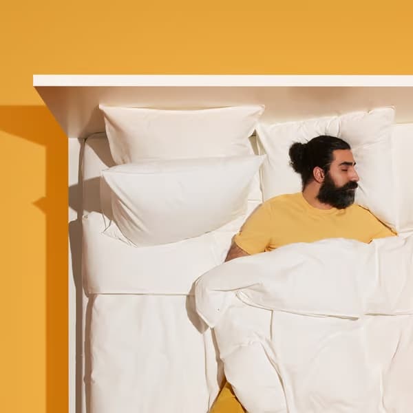 Persona recostada en una cama con ropa de cama blanca y almohadas, sobre un fondo amarillo.