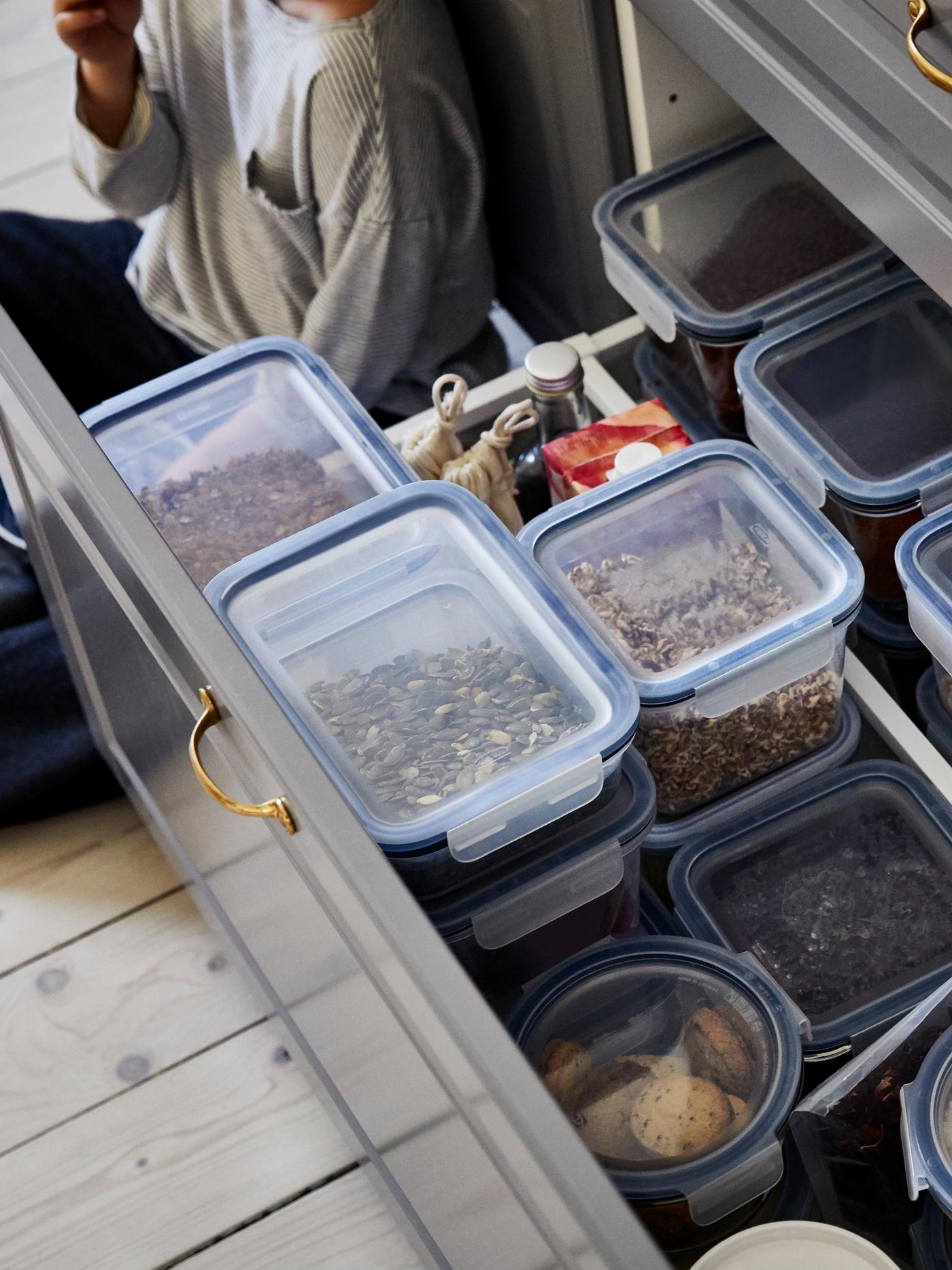 Persona seduta sul pavimento accanto a un cassetto aperto, pieno di contenitori per alimenti IKEA 365+ con semi, frutta secca e altri cibi.