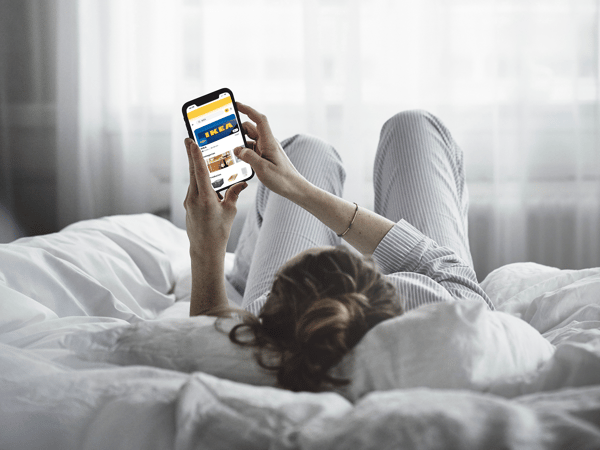 Persona tumbada en la cama con pijama consultando la web de IKEA en el móvil, en un ambiente relajado y acogedor, para la promoción de IKEA Cataluña y Glovo.