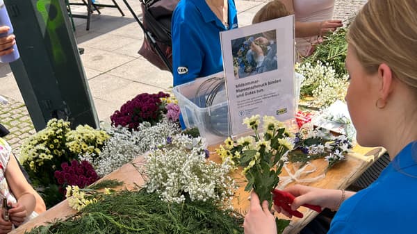 Personen binden Blumen und Grünes und schneiden Blumensträche mit einer Blumenschere. Es steht ein Schild mit der Überschrift 