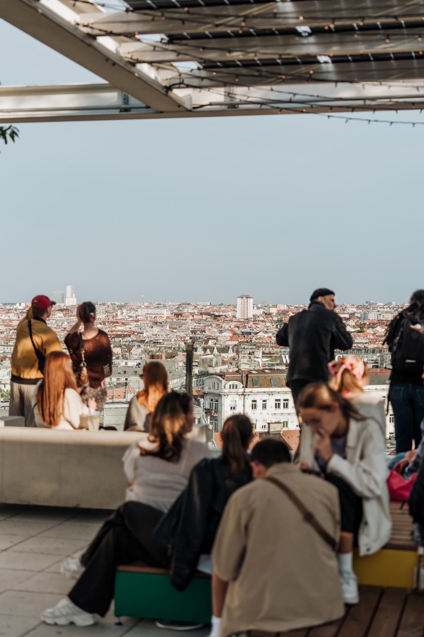 Personen die sitzend und stehend die Aussicht von der Dachterrasse genießen. Im Hintergrund ist der Ausblick auf Wien zu sehen.