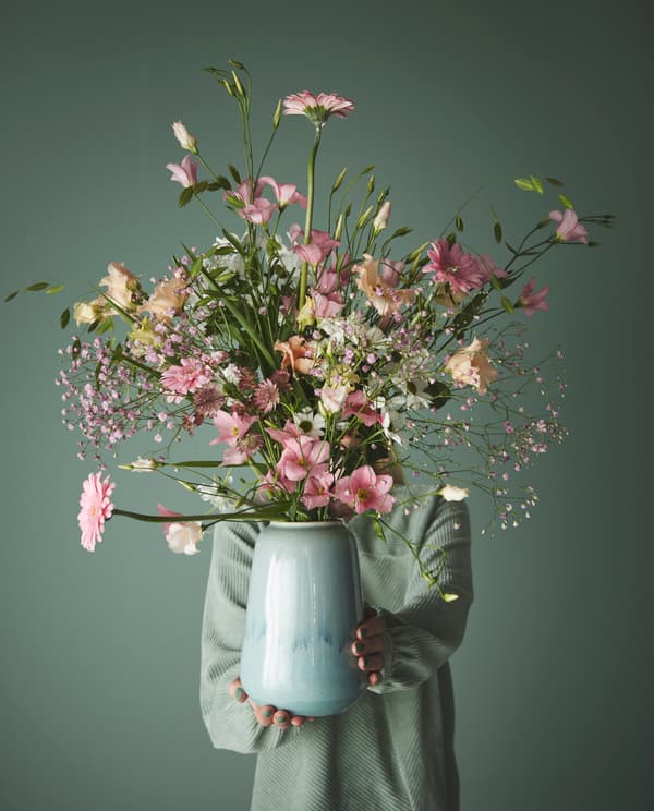 personne avec un pull vert portant un grand vase bleu rempli de grandes fleurs sur un fond vert
