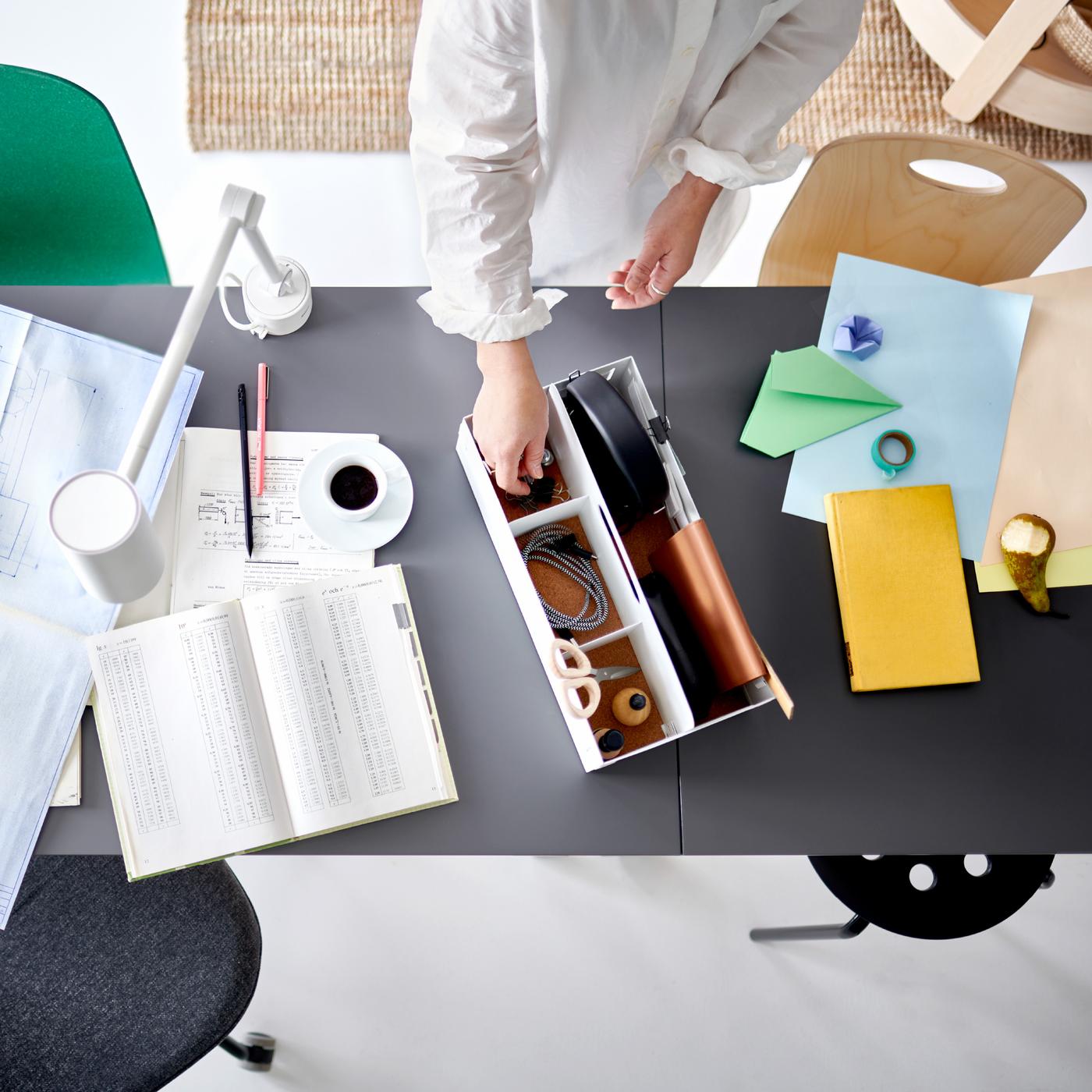 Personne debout contre un organiseur de bureau blanc posé sur deux plateaux de table jumelés, près de divers documents et d’une lampe de bureau blanche.