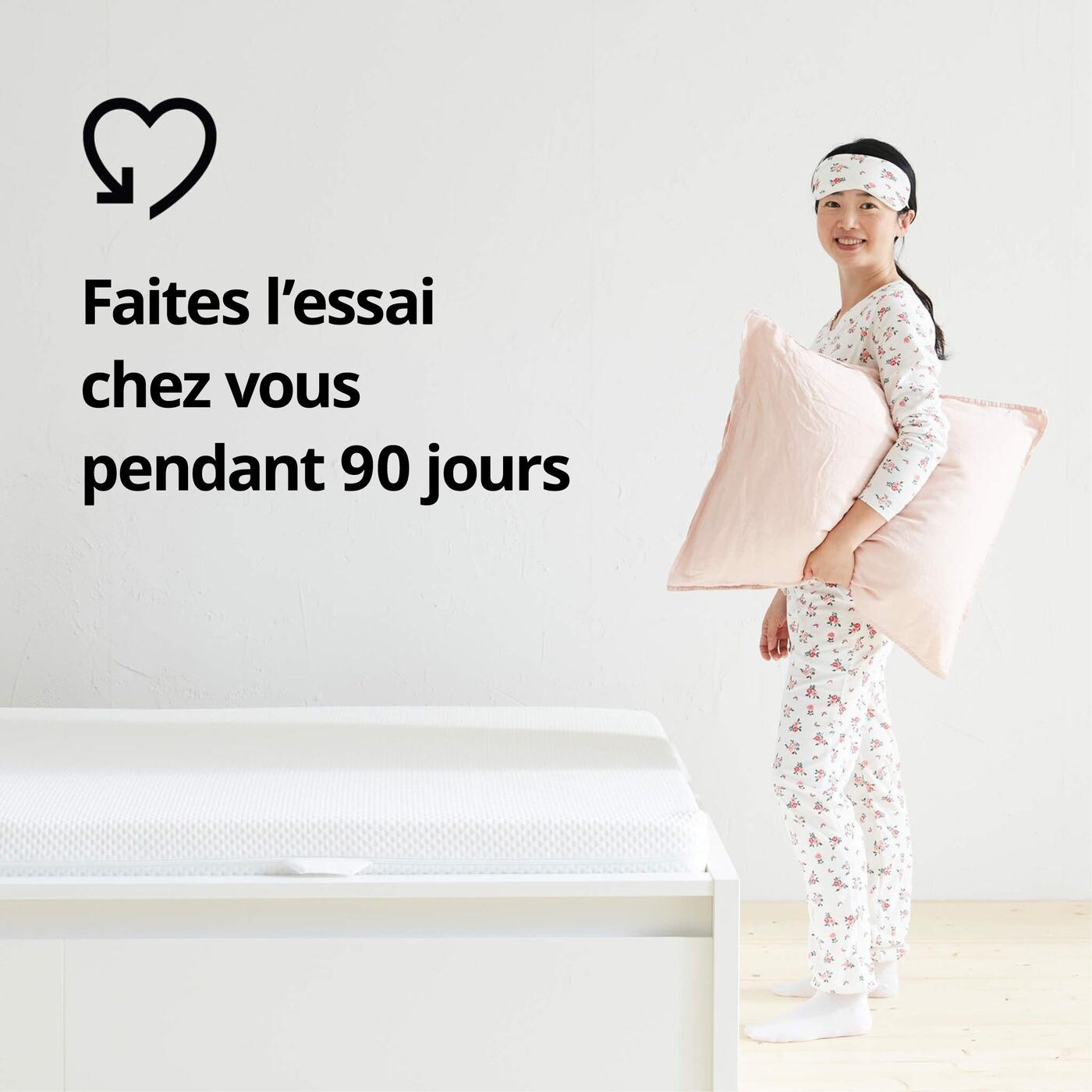 Personne en pyjama à côté d'un lit avec matelas, tenant un oreiller rose ; à gauche, texte « Faites l"essai chez vous pendant 90 jours » avec le pictogramme en forme de cœur.