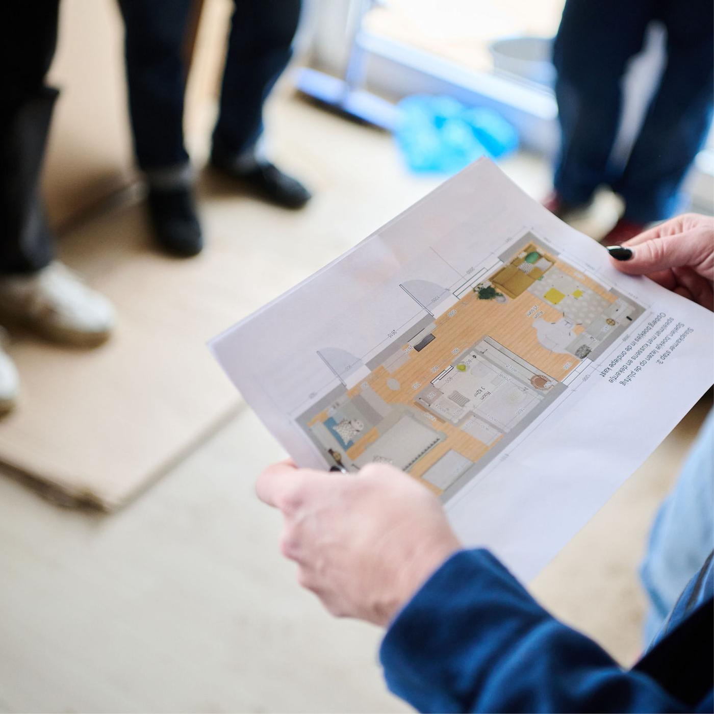Personne tient un plan d'aménagement intérieur avec d’autres personnes en arrière-plan dans une pièce vide.