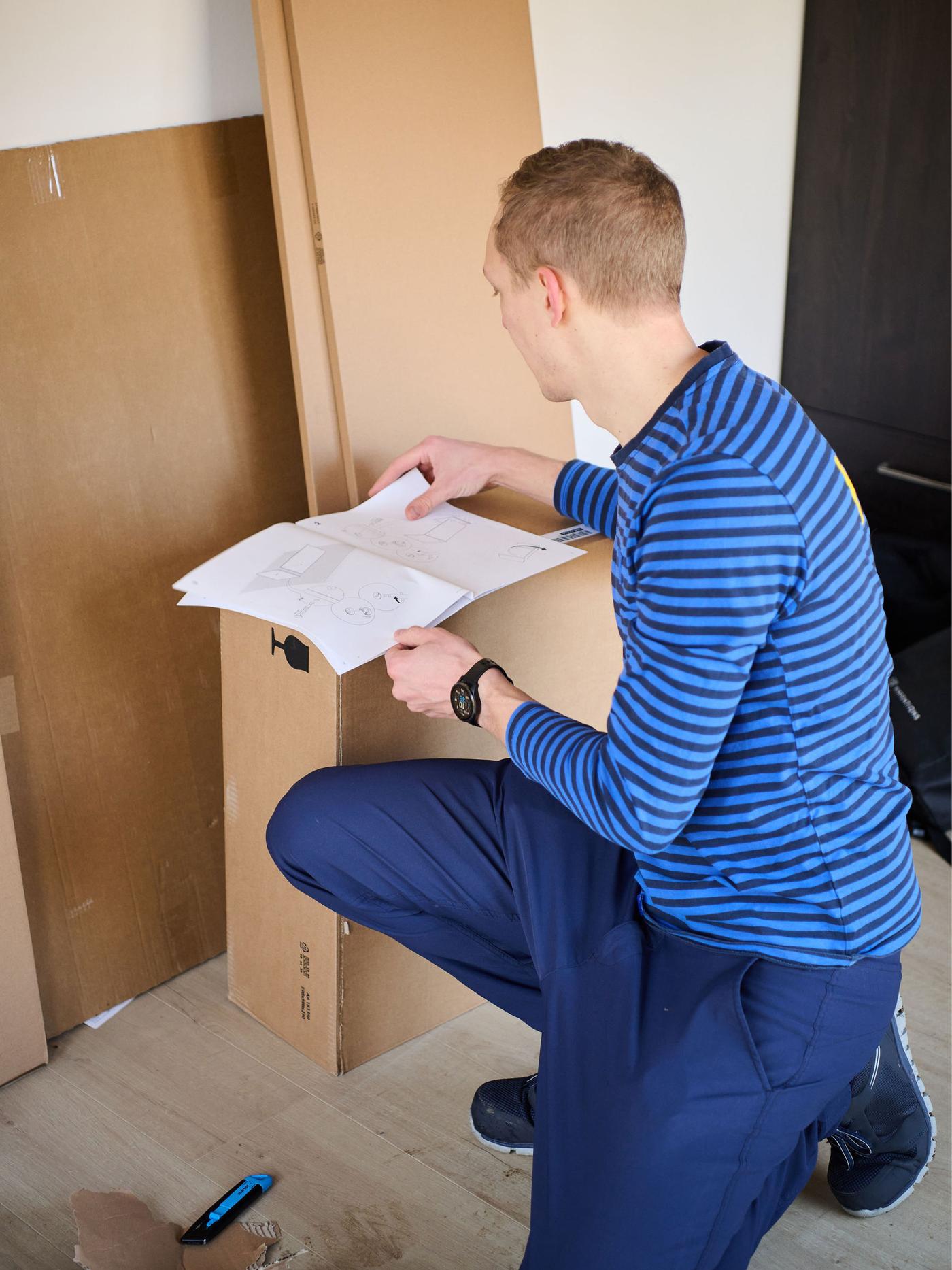 Persoon bekijkt een montagehandleiding naast een kartonnen doos en panelen op een houten vloer.