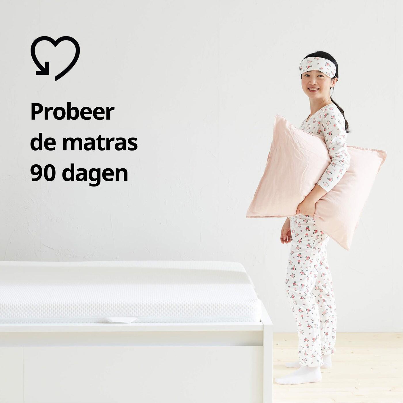 Persoon in pyjama naast bed met matras, houdt roze kussen vast; links staat de tekst "Probeer de matras 90 dagen" met hartvormig symbool.