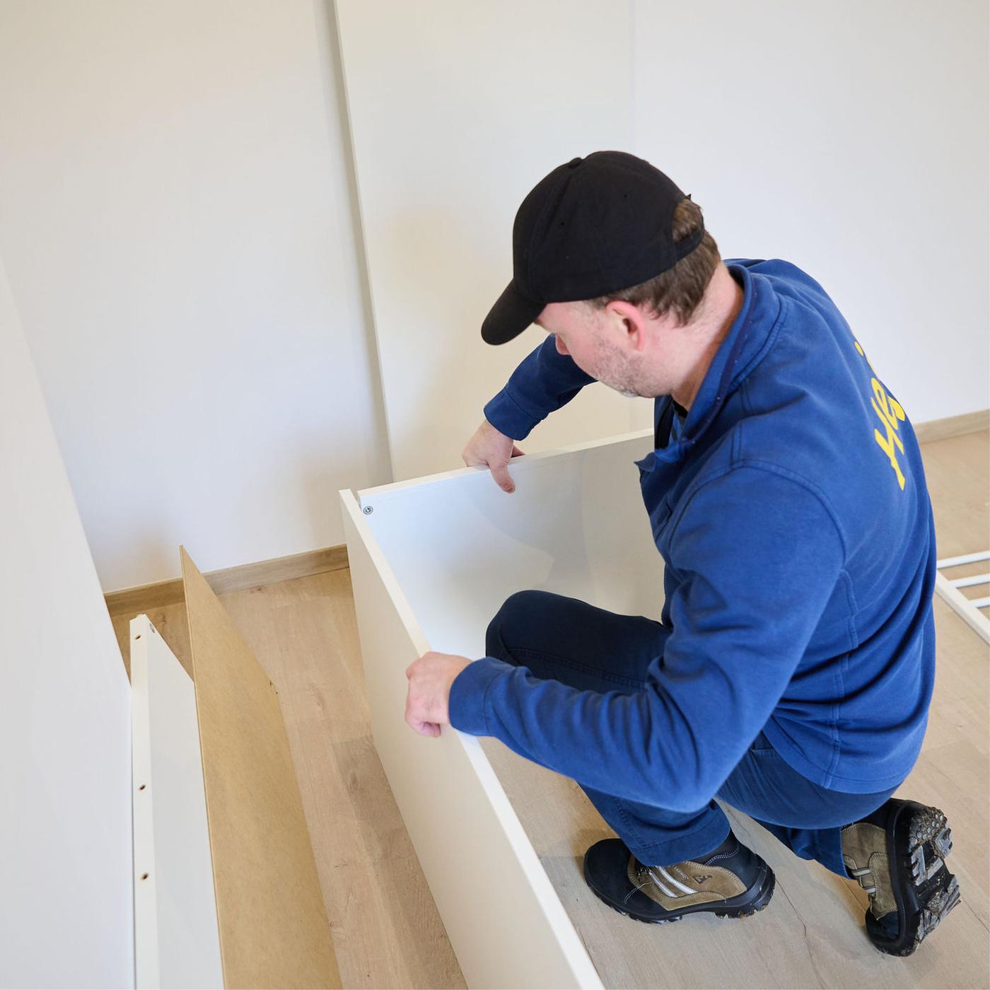 Persoon monteert een witte kastpaneel op een houten vloer in een lege kamer.