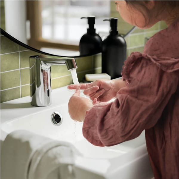 Persoon wast de handen onder een stromende kraan aan een moderne wastafel. Zeepdispensers en groene tegels zorgen voor een frisse uitstraling.