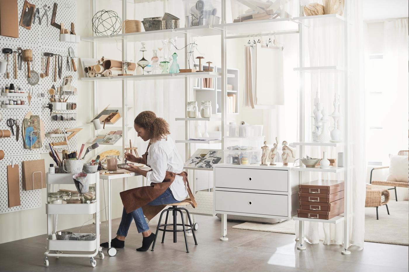 Persoon werkt aan bureau in lichte werkruimte met rekken, gereedschap en decoratieve objecten rondom.