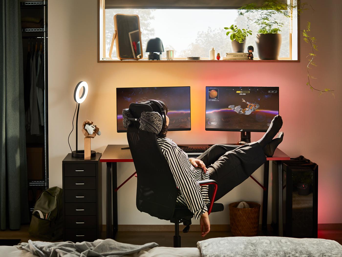 Pertsona bat HUVUDSPELARE aulki beltz batean etzanda dago, bi pantailen aurrean eta hankak gaming-mahai baten gainean dituela.