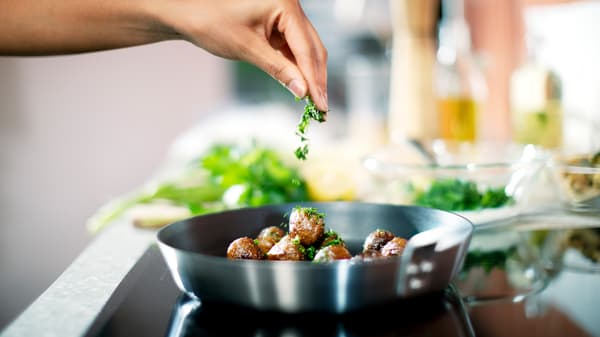 Pessoa polvilha ervas aromáticas picadas sobre almôndegas de proteína vegetal HUVUDROLL numa frigideira IKEA 365+ numa placa.