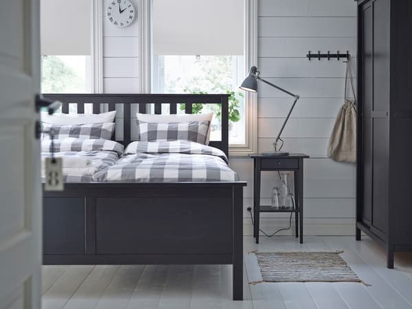 Petite chambre meublée d’un cadre de lit IKEA HEMNES en brun-noir avec literie à carreaux gris clair et blancs.