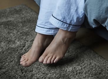 Peus descalços sobre una catifa tova de color gris, just al costat del llit, amb pantalons de pijama blancs.