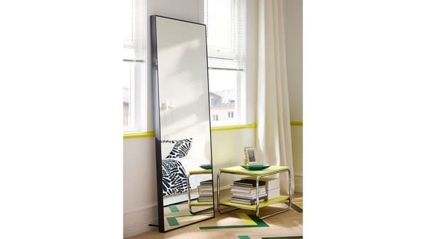 Photo of a black HOVET mirror leaning against a window with a white curtain next to a small yellow table with a picture frame, dish, and books on it. 