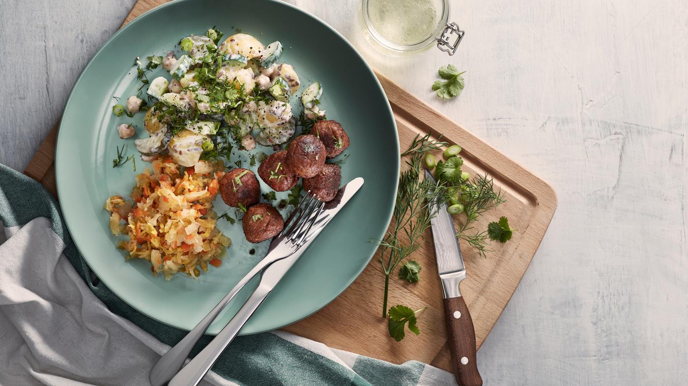 Piatto turchese con polpette vegetali, insalata di patate e kimchi su un tagliere, accanto a un coltello, erbe aromatiche e uno strofinaccio.