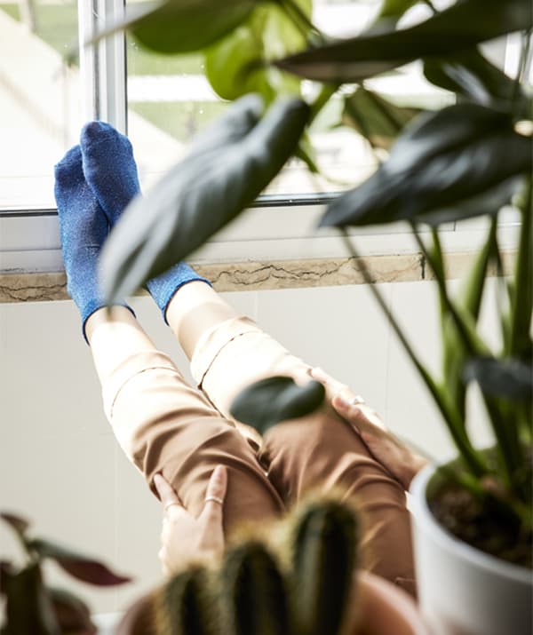 Picioarele unei femei sprijinite de pervaz, văzute printre plantele din ghivece.