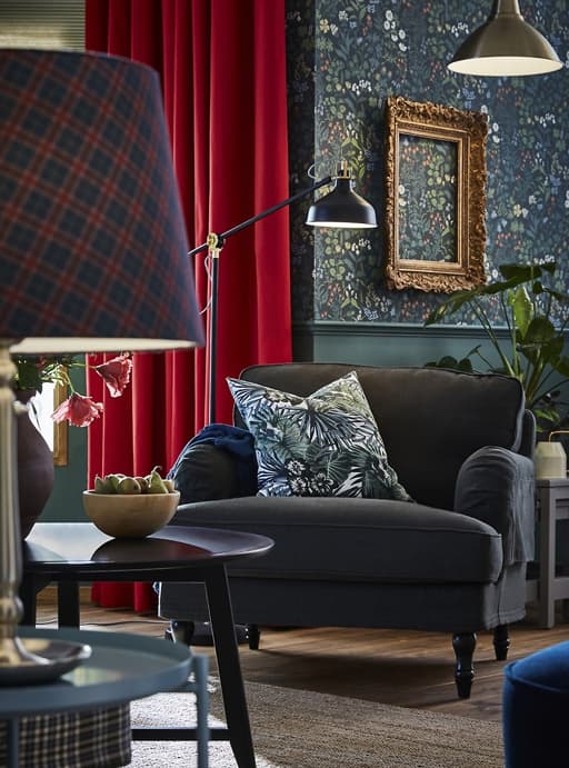 Picture of a dark green armchair with a floral cushion on it and a tartan lamp in the foreground on a side table.