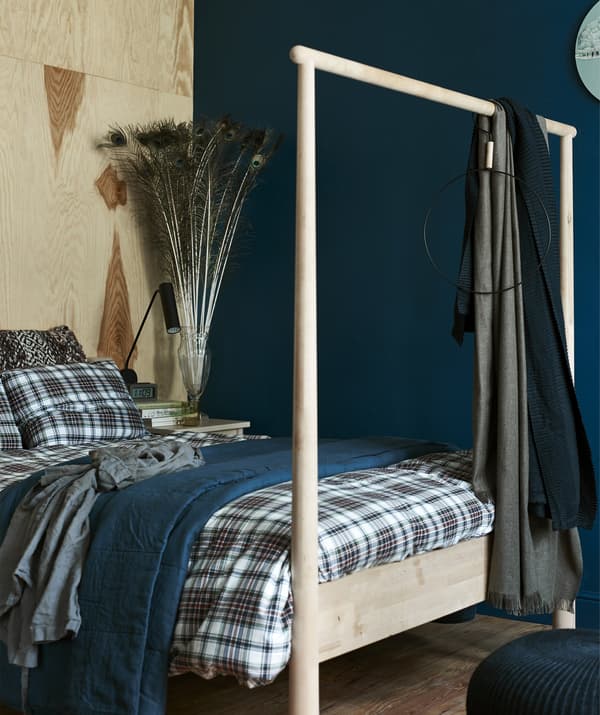 Picture of large wooden bedframe with layered bedding and large display of peacock feathers in a vase in the corner.