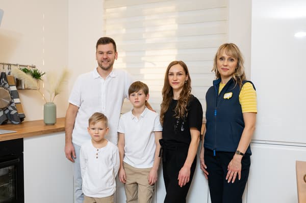 Pieci cilvēki stāv gaišā, modernā virtuvē – divi pieaugušie, divi zēni un IKEA darbiniece. Ģimene pozē kopā ar dizaineri pie baltām virtuves mēbelēm un loga ar žalūzijām.