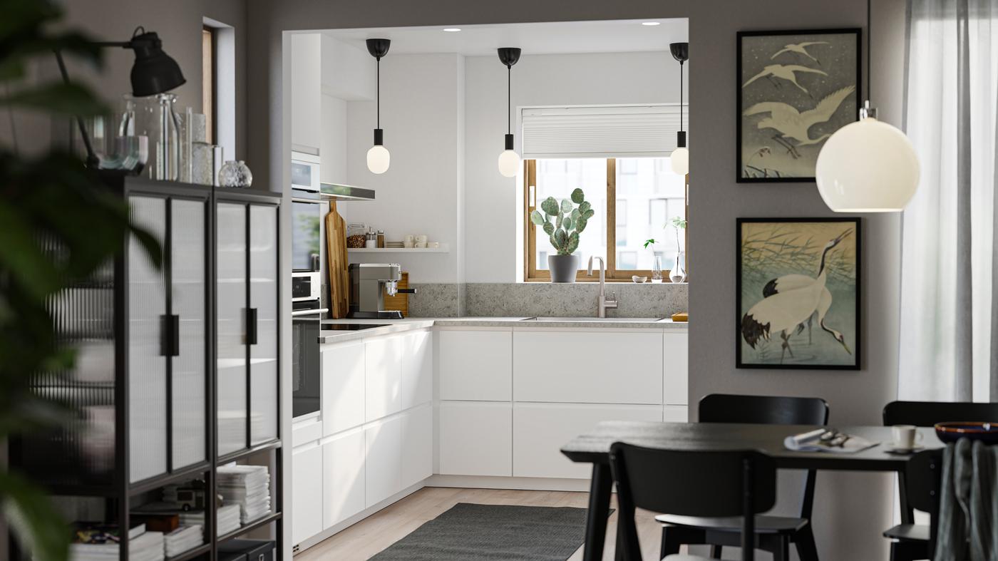 Pieni keittiö, jossa on sileät valkoiset VOXTORP-ovet, valkoiset seinät, harmaa matto lattialla ja kolme pientä mustaa kattovalaisinta.