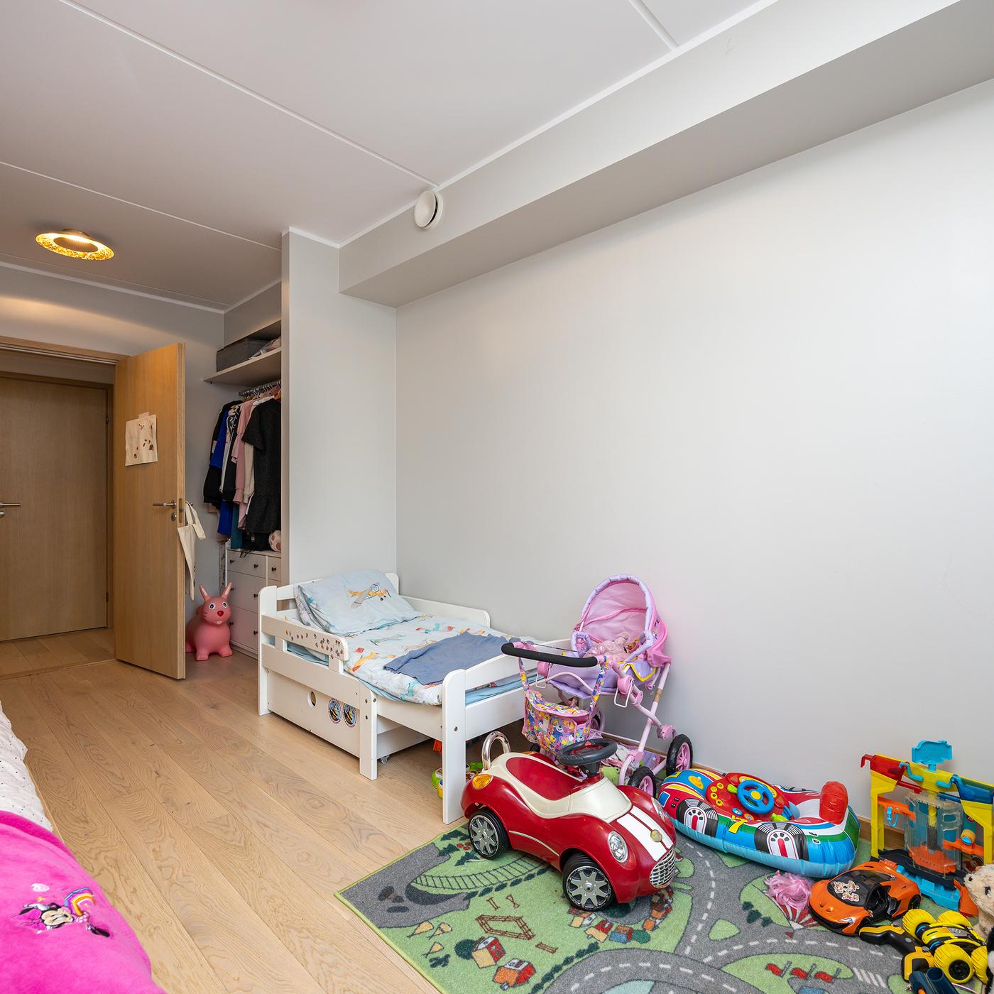 Piklik heledates toonides lastetuba. Esiplaanil paremas servas on valge kummut ja selle kohal ripuvad riidepuutorul värvilised riided. Tagapool on voodid.